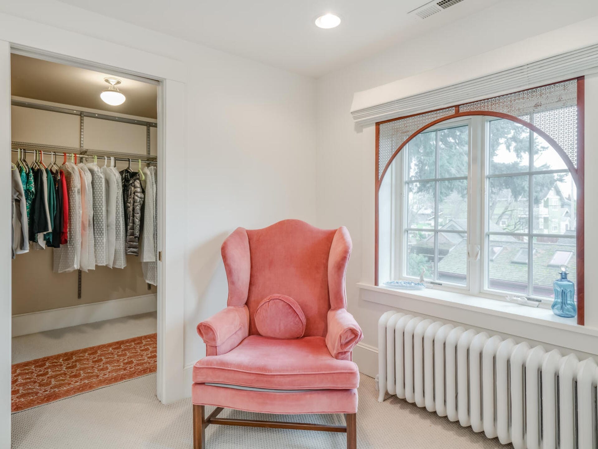 A cozy room features a pink upholstered armchair near a large arched window with a view of trees. To the left, a walk-in closet displays neatly hung clothes. A radiator sits below the window, and a decorative bottle adorns the windowsill.