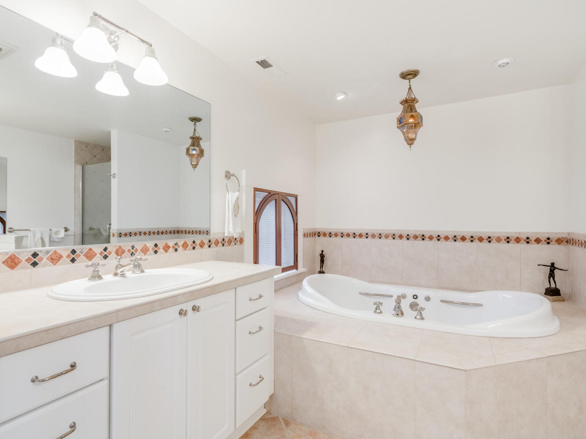 A clean, bright bathroom with a corner bathtub surrounded by beige tiles. A large mirror and double sink with white cabinets are on the left. Two ornate pendant lights hang overhead, and a small window adds natural light.