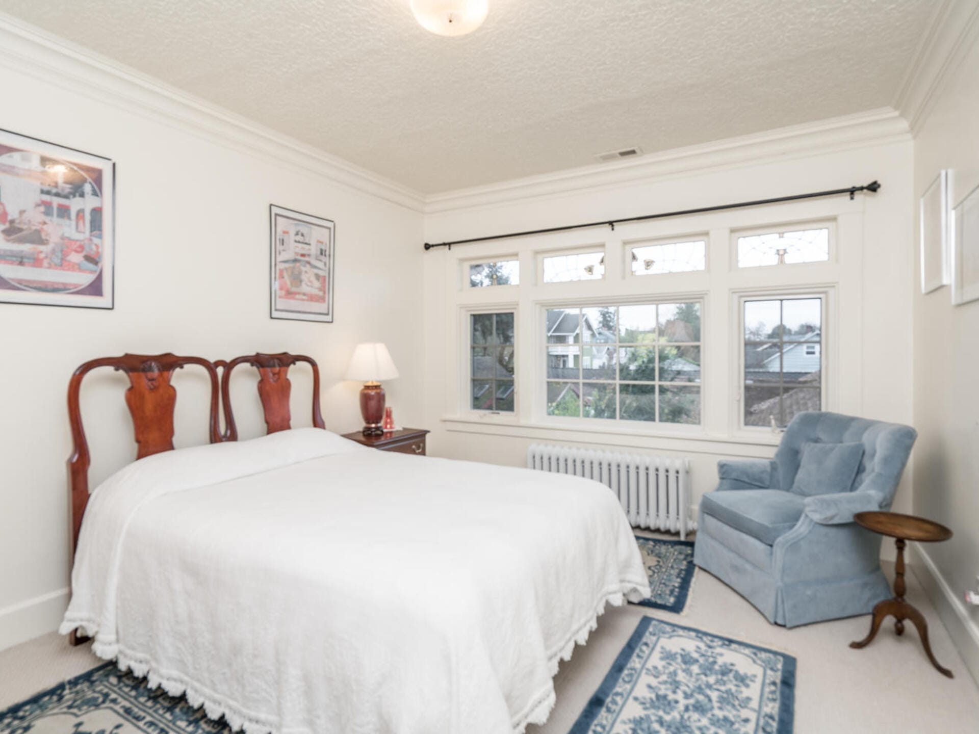 A cozy bedroom with a large bed covered in a white quilt. Two wooden chairs serve as a headboard. Theres a blue armchair and a small round table by the window. The room features wall art, a table lamp, and patterned rugs on a light floor.