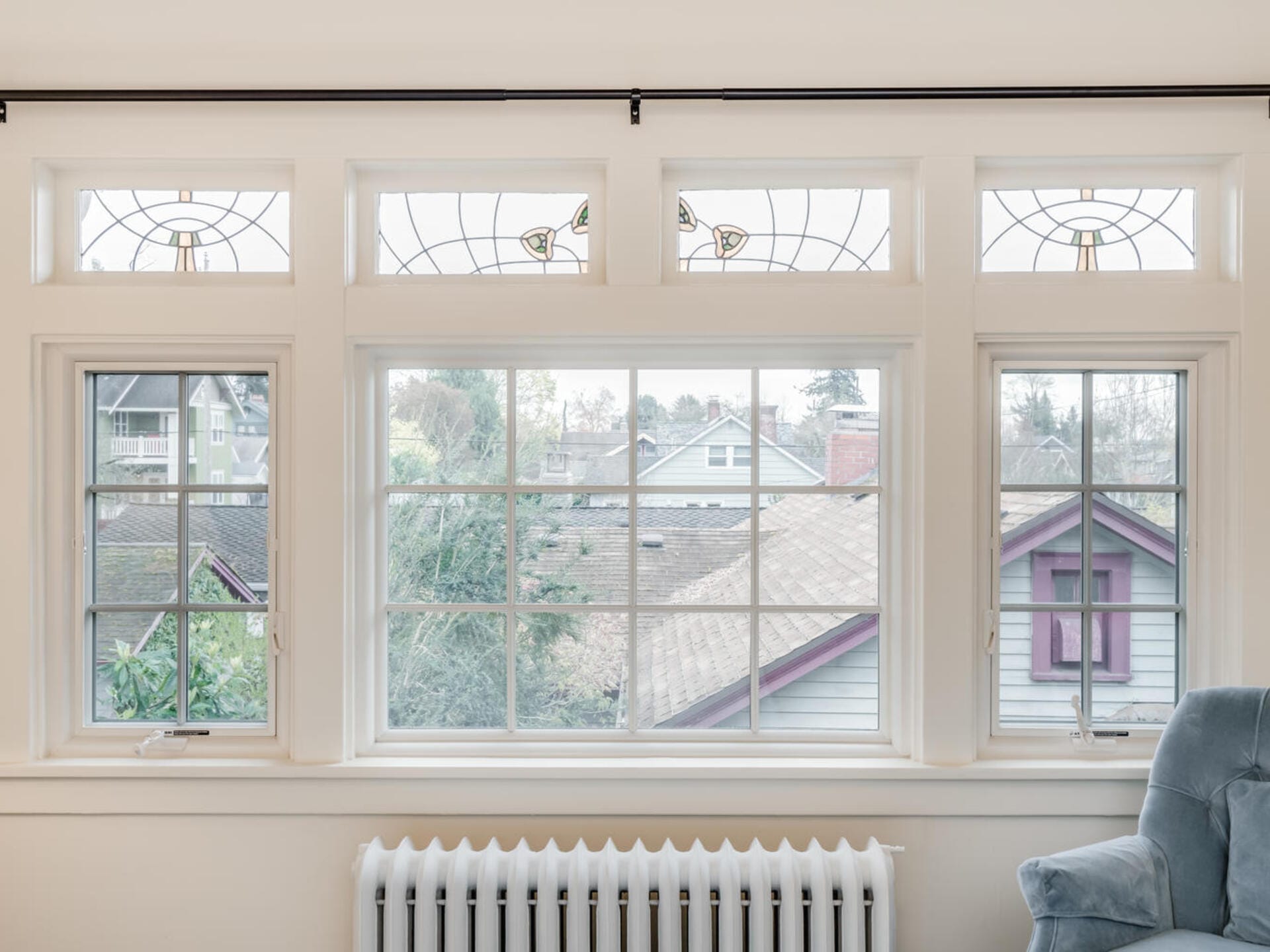 A large window with decorative stained glass panels at the top overlooks a residential neighborhood with houses and trees. Below the window, a white radiator is visible. A soft, light-colored chair sits to the right.