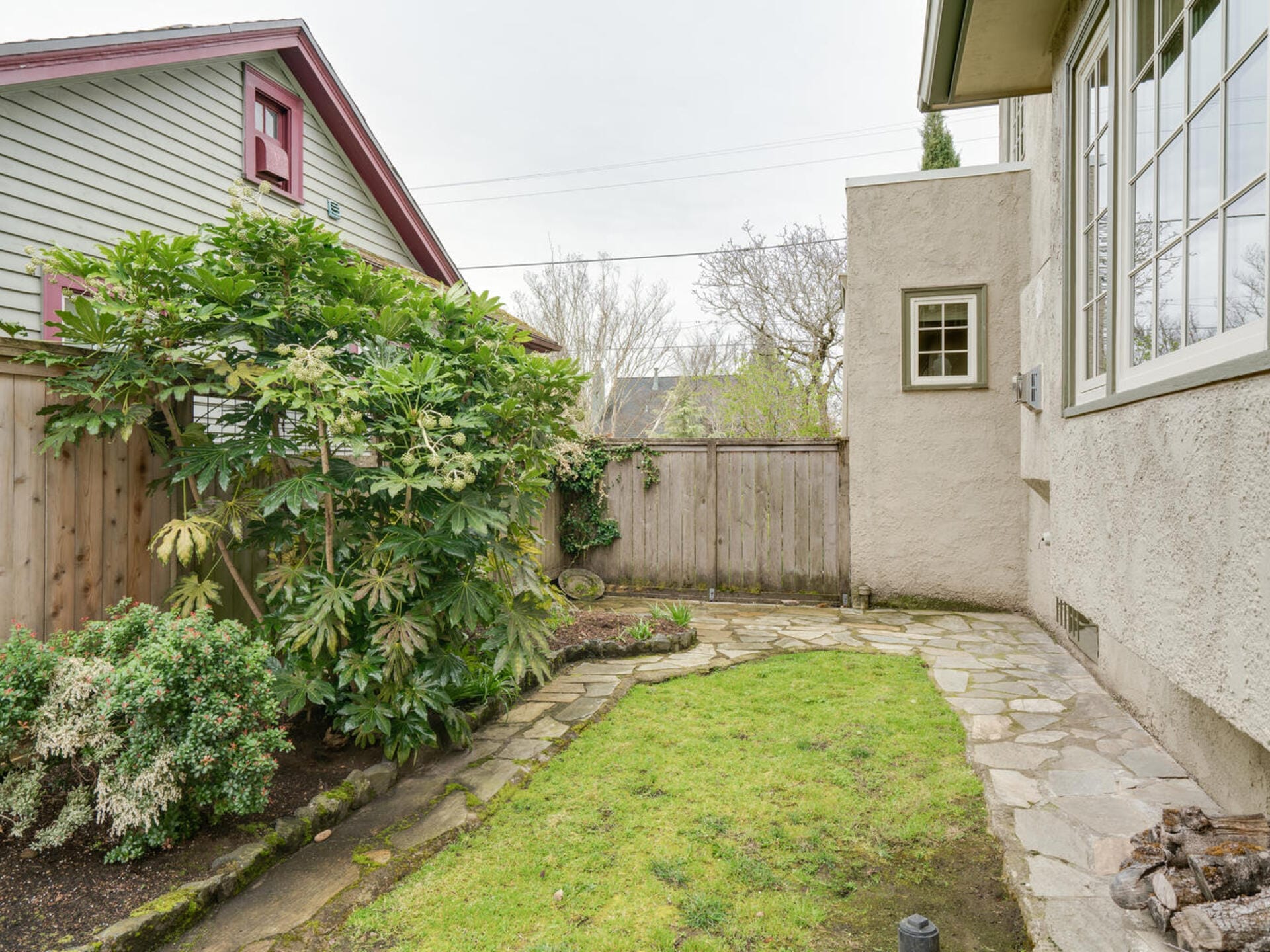 A small, fenced backyard with a narrow stone pathway and a patch of grass. Lush green shrubs and trees line the left side, while a neighboring house is partially visible. The right side features part of a beige house with large windows.