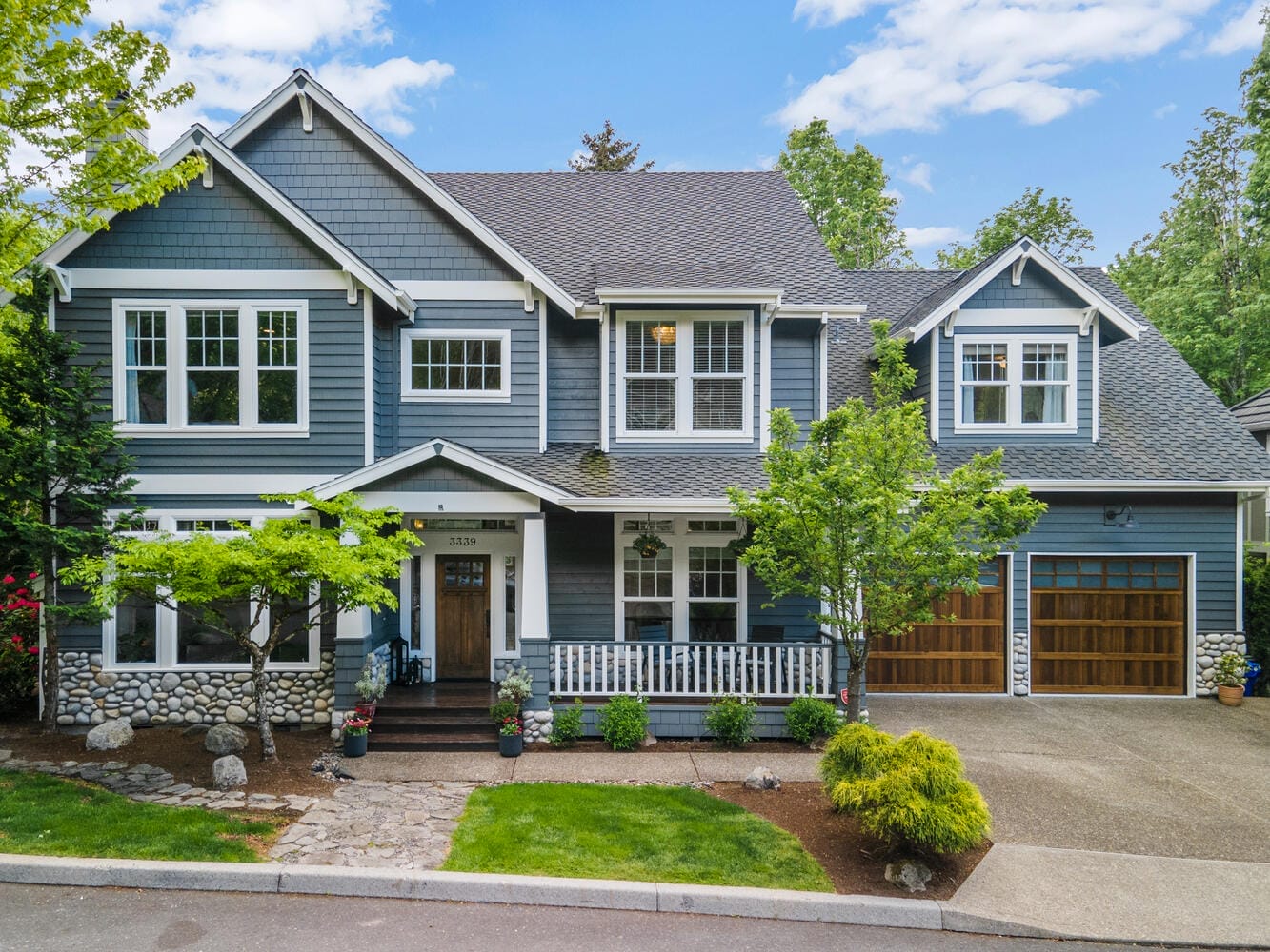 A large, two-story house with blue siding, white trim, and a stone facade. It has a front porch with white railings, two wooden garage doors, and is surrounded by green trees and landscaping. The sky is partly cloudy.