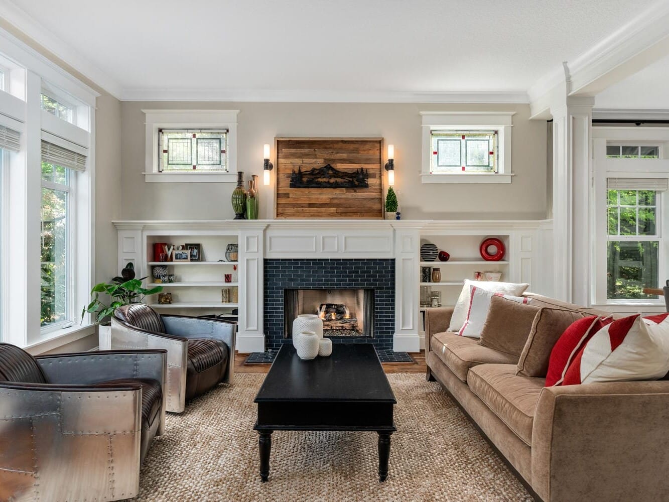 A cozy living room featuring a fireplace with a wooden mantle, surrounded by built-in shelves. Two leather chairs and a brown sofa with red and white cushions are arranged around a dark coffee table. Large windows let in natural light.