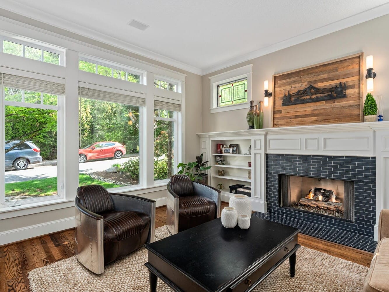 Cozy living room with hardwood floors, two leather chairs, a black coffee table, and a fireplace with decorative wood paneling. Large windows on the left overlook a garden and parked cars outside. Neutral-toned walls and built-in shelves complete the space.