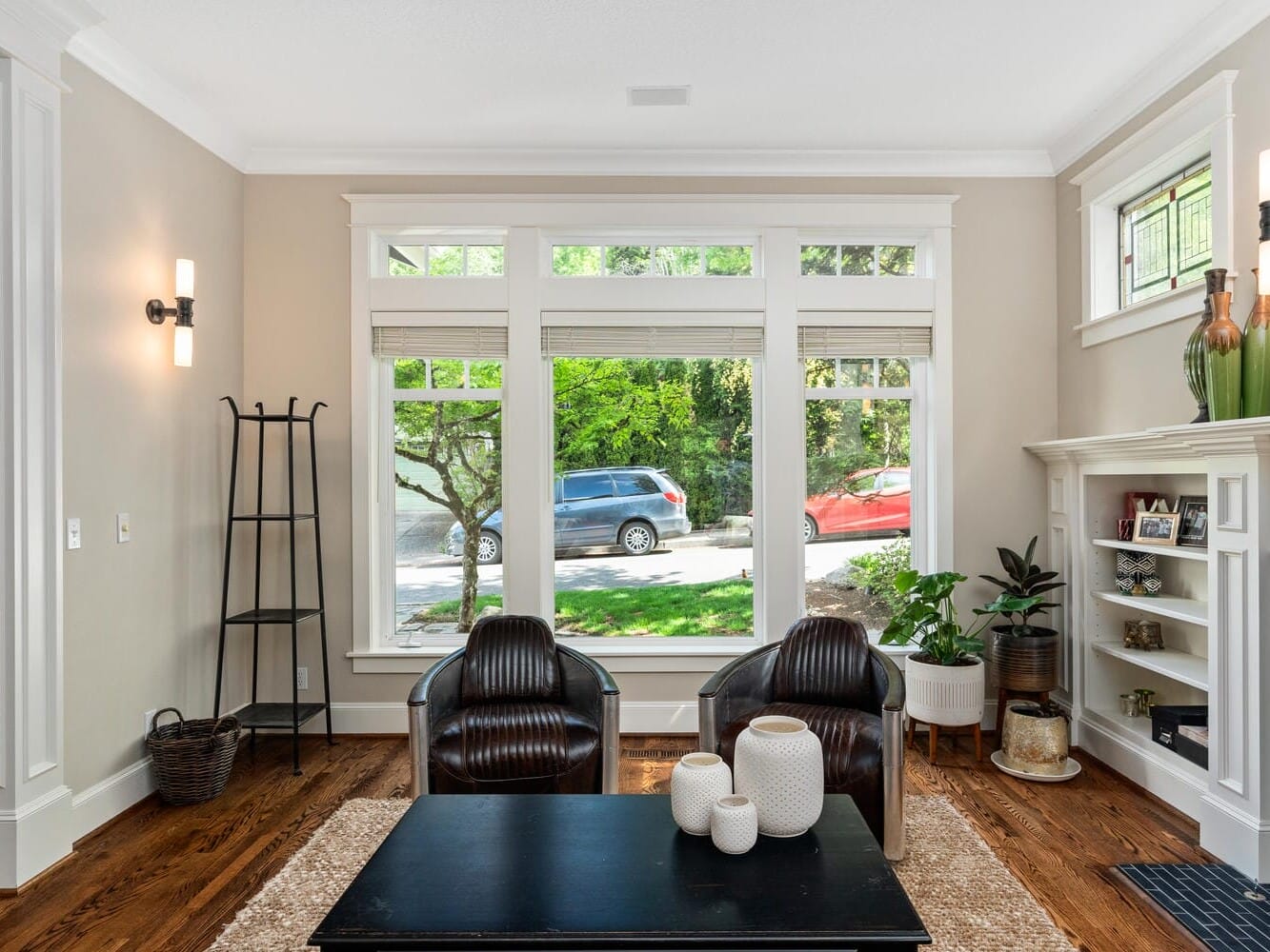 A cozy living room with two black leather chairs facing a black coffee table. Large windows showcase a view of green trees and parked cars. A fireplace with shelves, plants, and decorative items are on the right. Natural light fills the space.