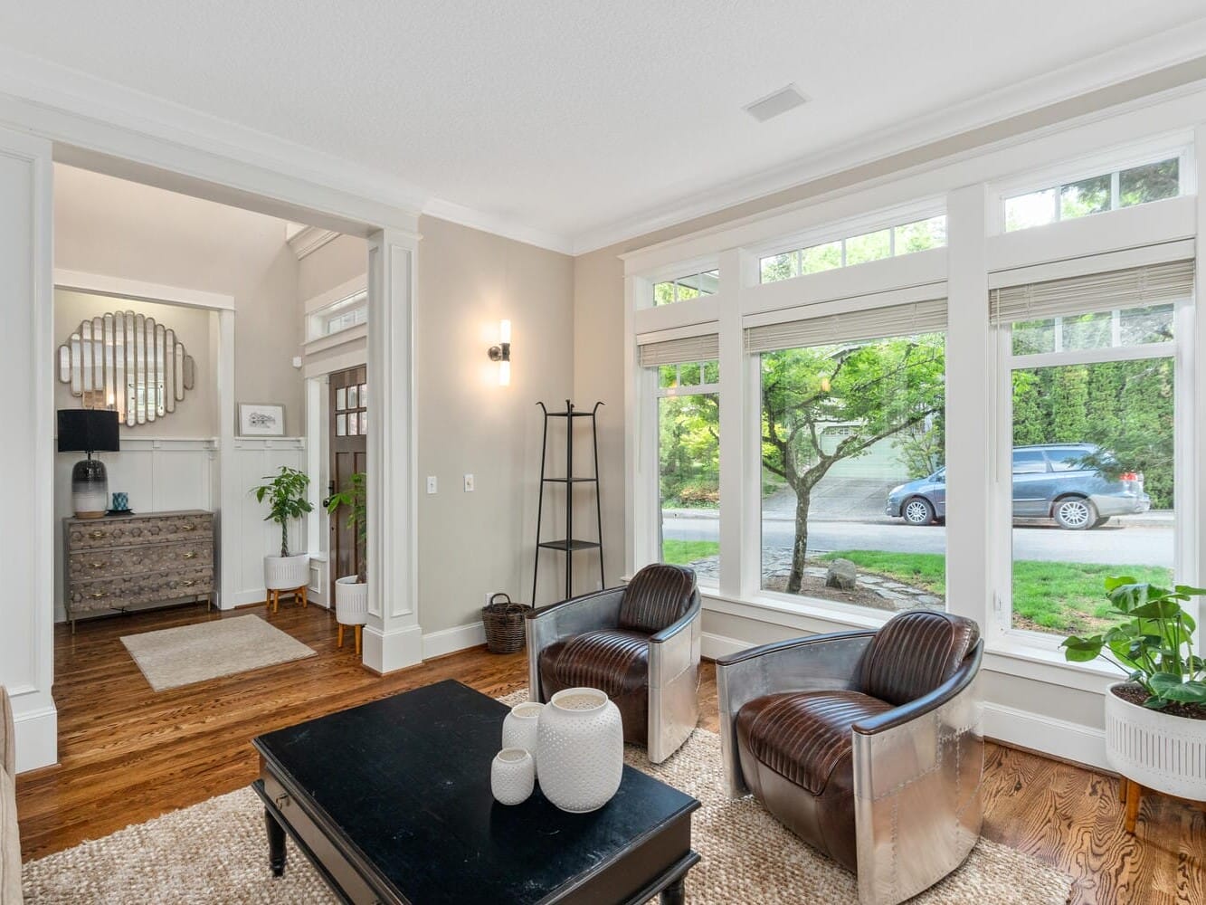 A modern living room with large windows showing a view of a car and greenery outside. The room features a black coffee table, two brown leather chairs with metallic accents, and potted plants. White walls and a hardwood floor complete the setting.