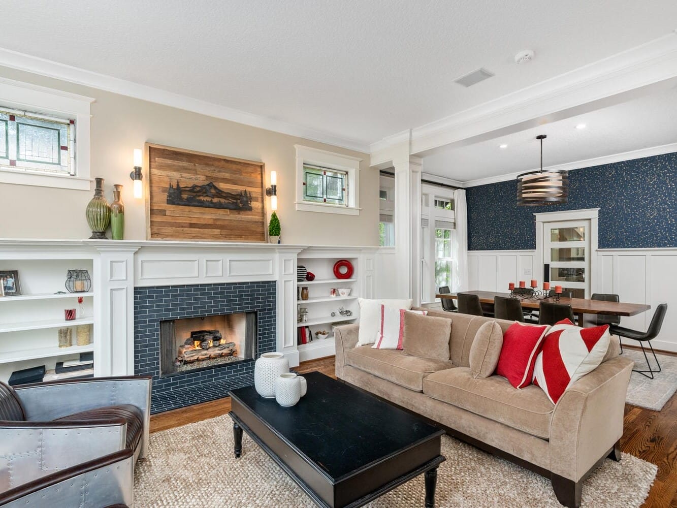 A cozy living room with a beige sofa adorned with red and white pillows. A fireplace with decorative shelves and art above it sits to the left. A wooden coffee table is centered on a textured rug. A dining area is visible in the background.