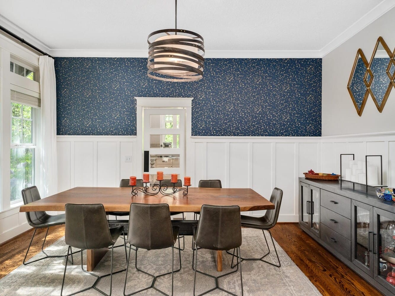 A dining room with a wood table, eight black chairs, and a decorative candle holder. The navy accent wall features gold speckles and diamond-shaped mirrors. A sideboard with decor items sits on the right, and large windows allow natural light in.