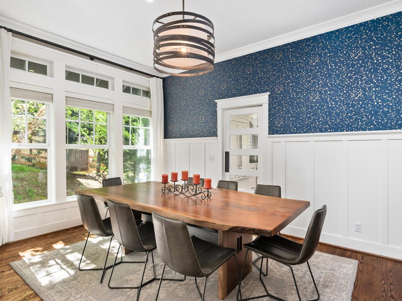 Bright dining room with a large wooden table, black chairs, and a centerpiece with red candles. The walls feature white wainscoting and a blue speckled upper section. Large windows let in natural light, highlighting the hardwood floor and gray rug.