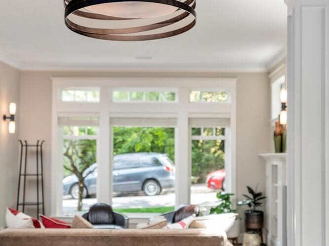 A modern living room with a metal light fixture hanging above a table. On the table is a candle holder with six red candles. The background features a sofa with red and beige cushions facing large windows revealing a view of trees and a parked car.