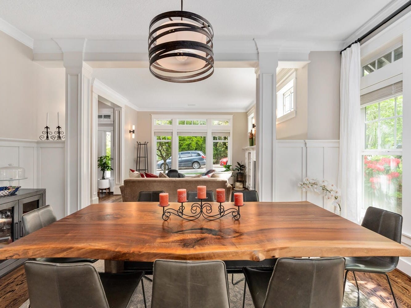 A modern dining room features a wooden table with six chairs and a candelabra centerpiece. In the background, a living room with a sofa and large windows offers a view of a green garden. A decorative chandelier hangs above.