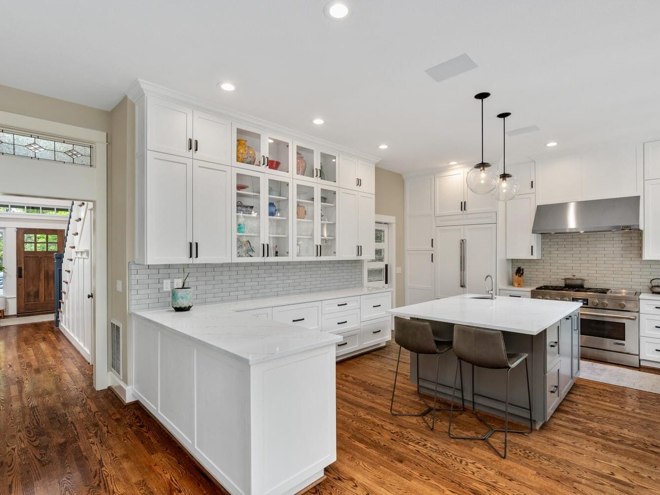 A spacious kitchen with white cabinets, a large island with two barstools, and hardwood floors. Modern appliances include a stainless steel stove and range hood. Recessed lighting and pendant lights illuminate the space. A wooden front door is visible.