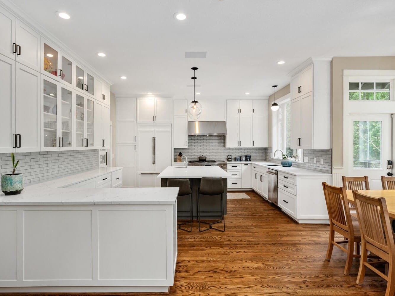 A modern kitchen with white cabinets, a large island with two chairs, stainless steel appliances, a dining table with wooden chairs, and hardwood flooring. Pendant lights hang from the ceiling, and windows let in natural light.