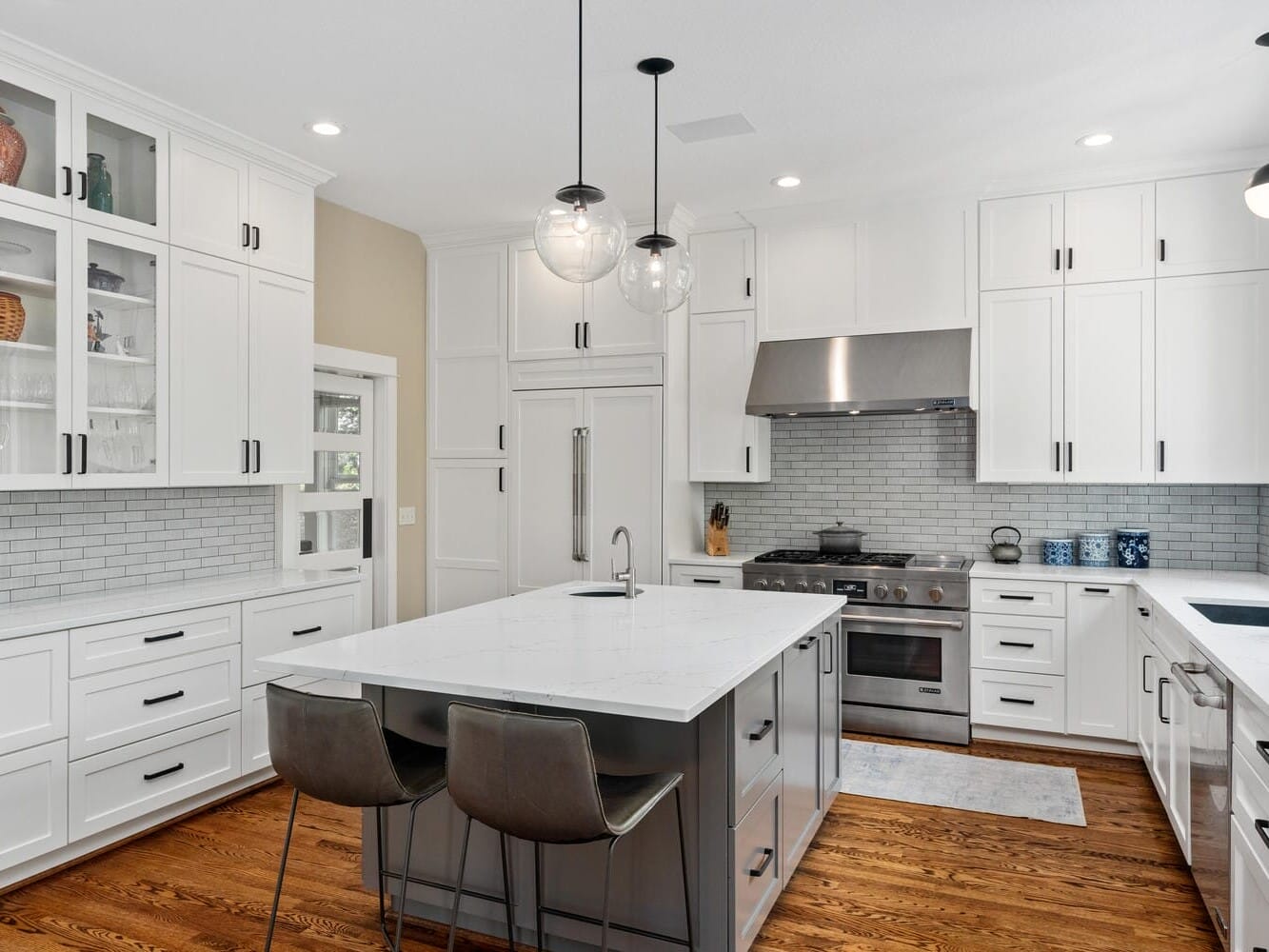 A modern kitchen featuring white cabinets, a large island with a marble countertop and two gray barstools. Stainless steel appliances include a stove and range hood. Pendant lights hang from the ceiling, and a subway tile backsplash completes the look.