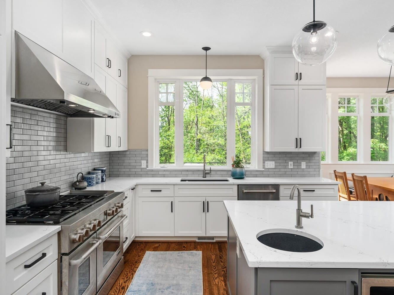 A modern kitchen with white cabinets, a stainless steel range hood, and a marble island with a sink. Two pendant lights hang above the island. Large windows provide natural light, overlooking greenery. A dining table is visible in the adjoining room.