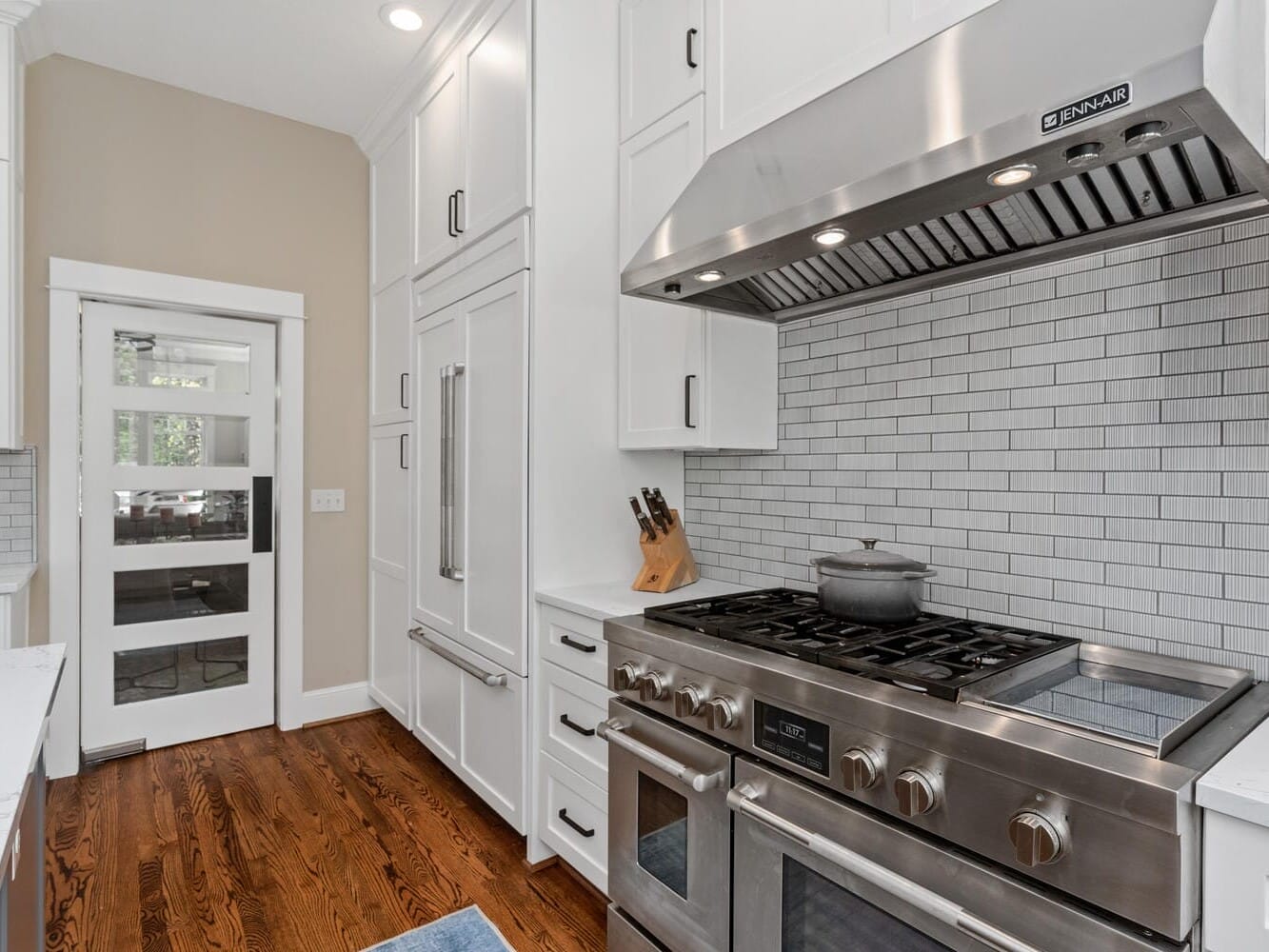 A modern kitchen with white cabinets, a stainless steel gas stove, and a matching vent hood. The backsplash features light gray tiles, and a pot sits on the stove. Wooden flooring and a glass door leading to another room are visible.