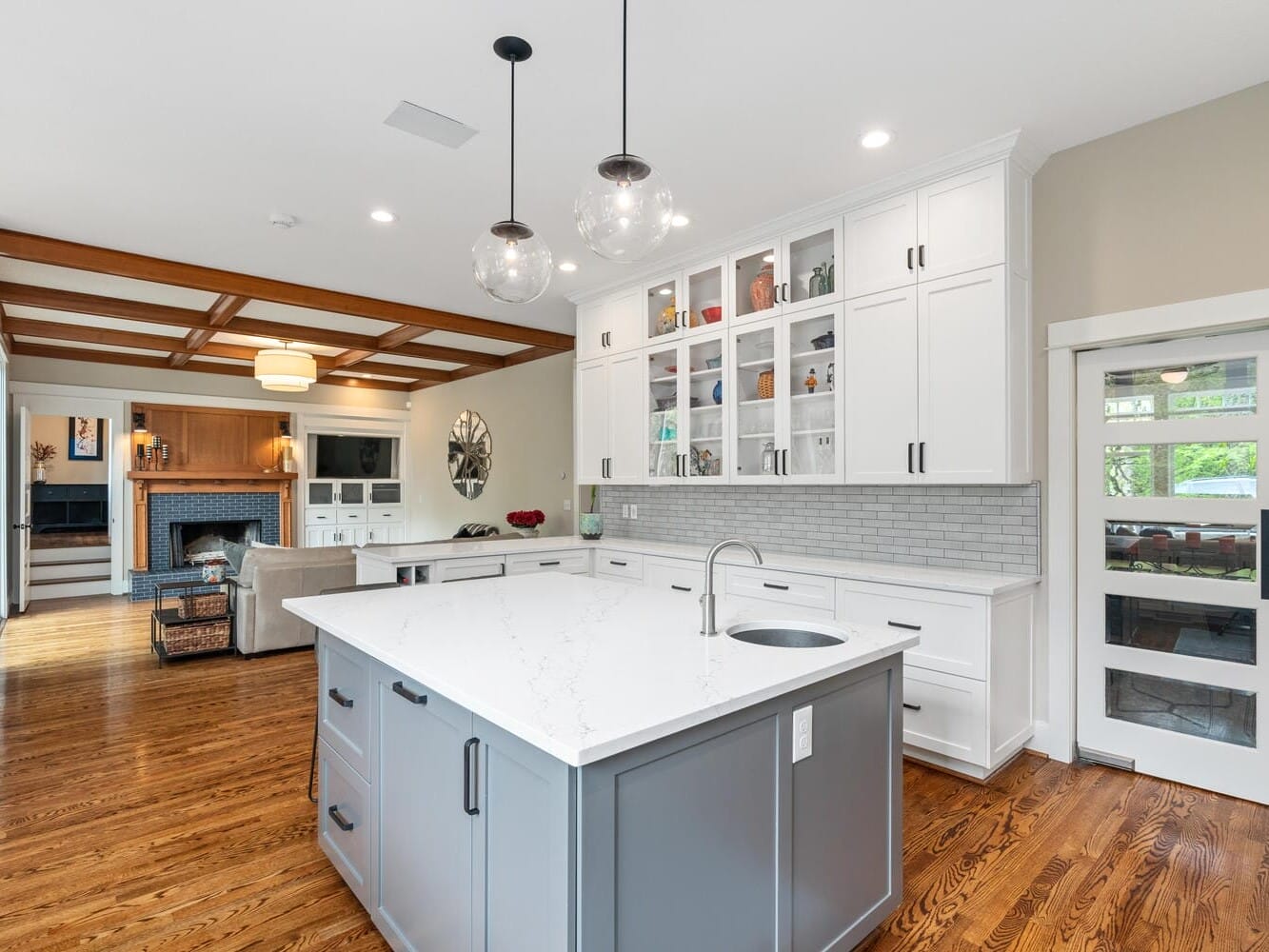 A spacious kitchen with a large white island, gray base, and built-in sink. White cabinets with glass doors line the walls. Open living space features hardwood floors and a cozy seating area with a fireplace. Pendant lights hang above the island.