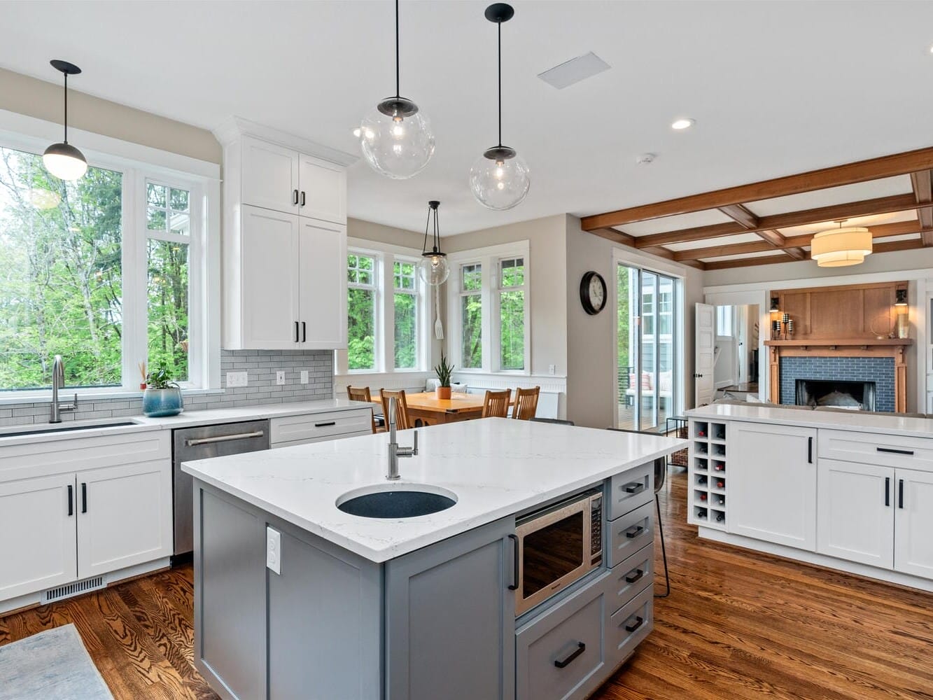 A bright, modern kitchen featuring a large island with a sink and oven, white cabinetry, and pendant lights. There is a dining area with a wooden table and chairs, large windows, and a cozy living space with a fireplace in the background.