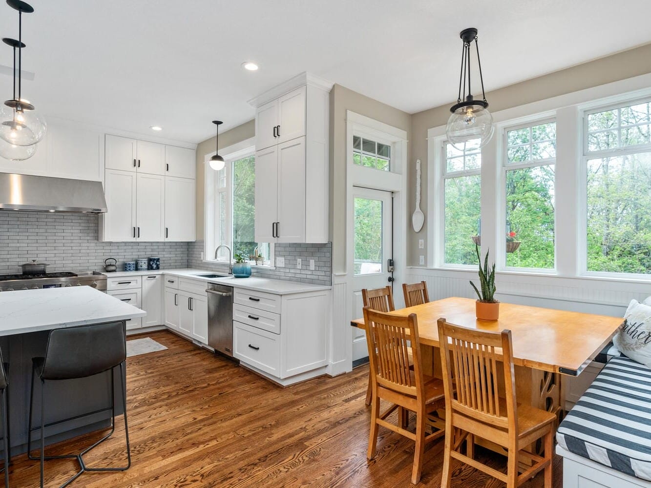 A modern kitchen with white cabinets and stainless steel appliances. An island with black chairs, pendant lights above, and a dining area with a wooden table and chairs. Large windows allow natural light to fill the space.