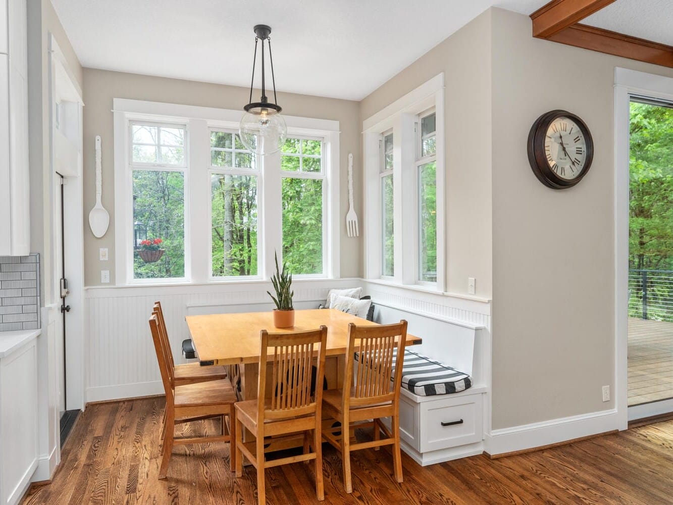 A cozy dining nook with a wooden table and four chairs beside a large window. A bench with a striped cushion is against the wall, and a modern pendant light hangs above. The area has a view of a lush green forest through the windows.