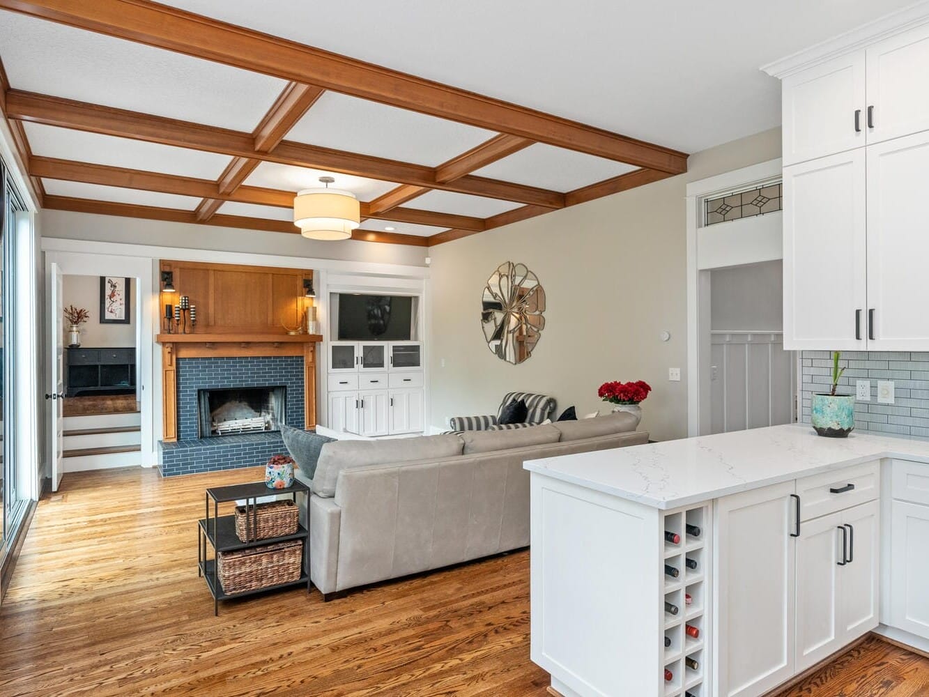 A cozy living room with a wooden coffered ceiling, beige walls, and hardwood floors. The room features a beige sectional sofa, a brick fireplace with a wooden mantel, built-in shelves, a wall mirror, and a white kitchen island with a wine rack.