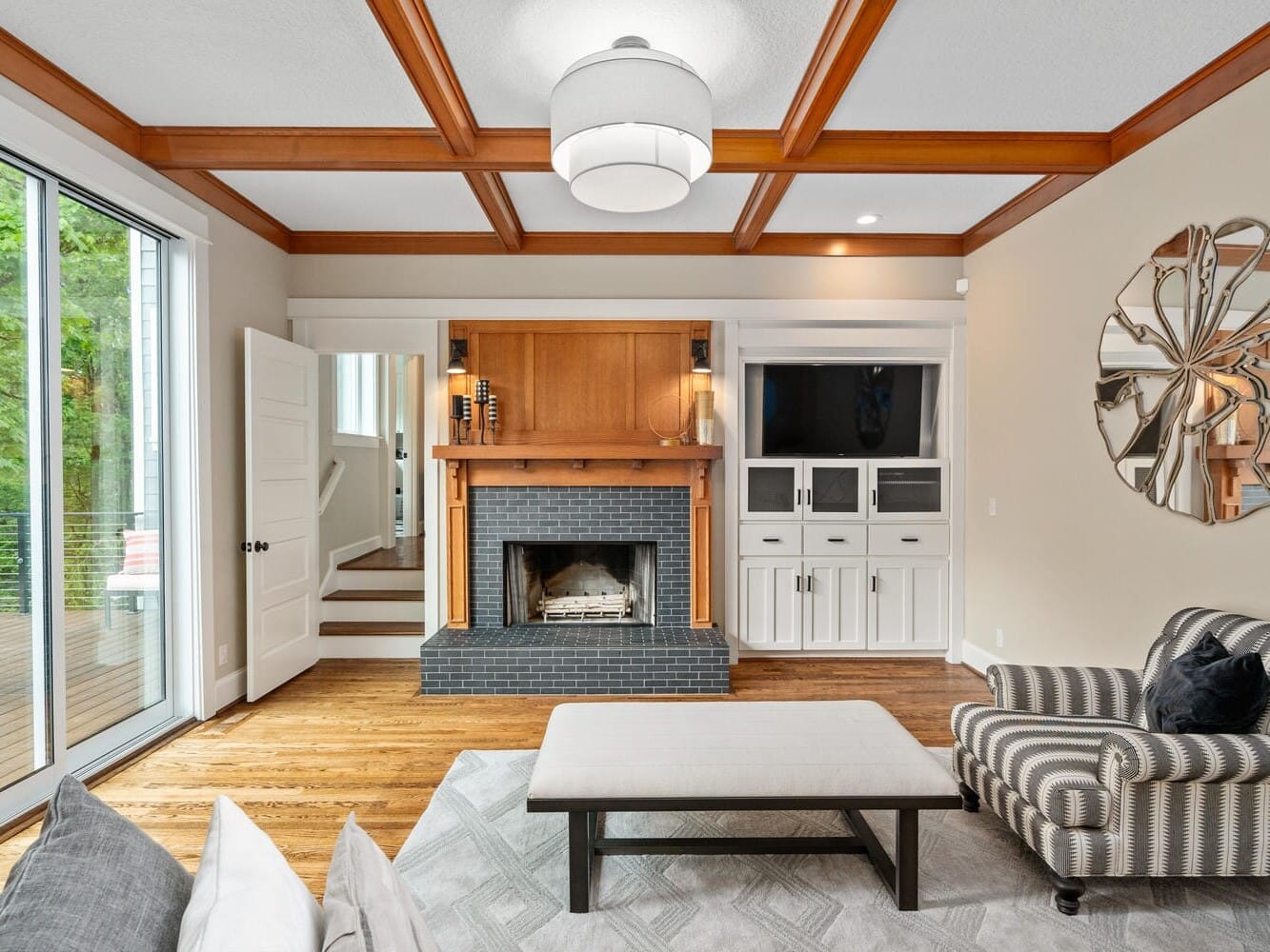 A cozy living room with a fireplace and a black mantle. Two built-in shelves with cabinets flanking the fireplace hold a TV and decor. A striped armchair, cushioned bench, and a geometric mirror enhance the warm wood and neutral-toned space. Large windows open to a deck.