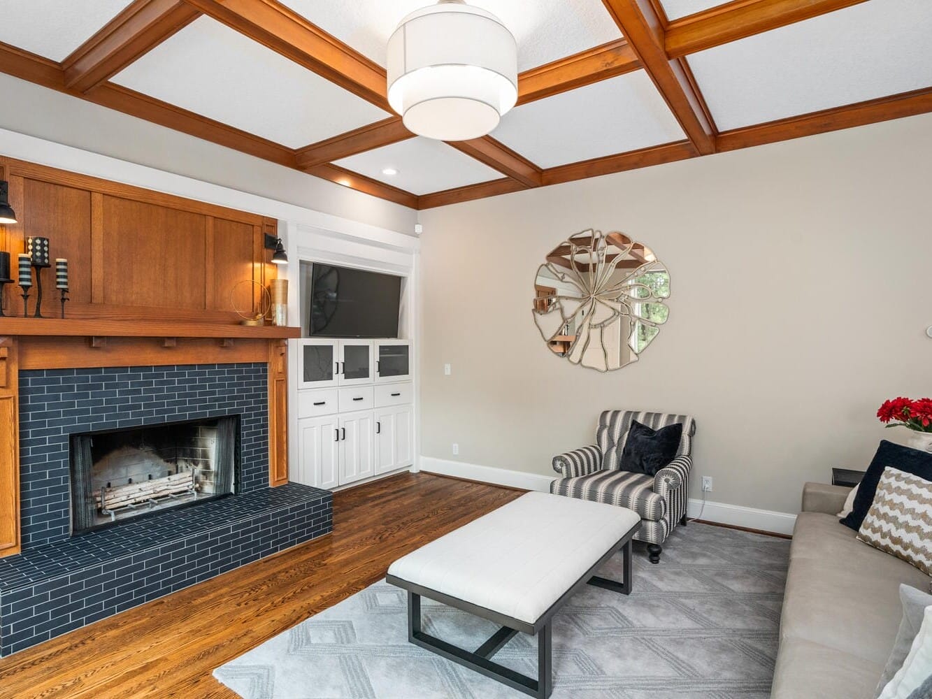 A cozy living room featuring a modern black fireplace, a wooden mantel, a striped armchair, a light gray sofa with patterned pillows, a rectangular coffee table, and a circular decorative mirror on the wall. The ceiling has wooden beams.