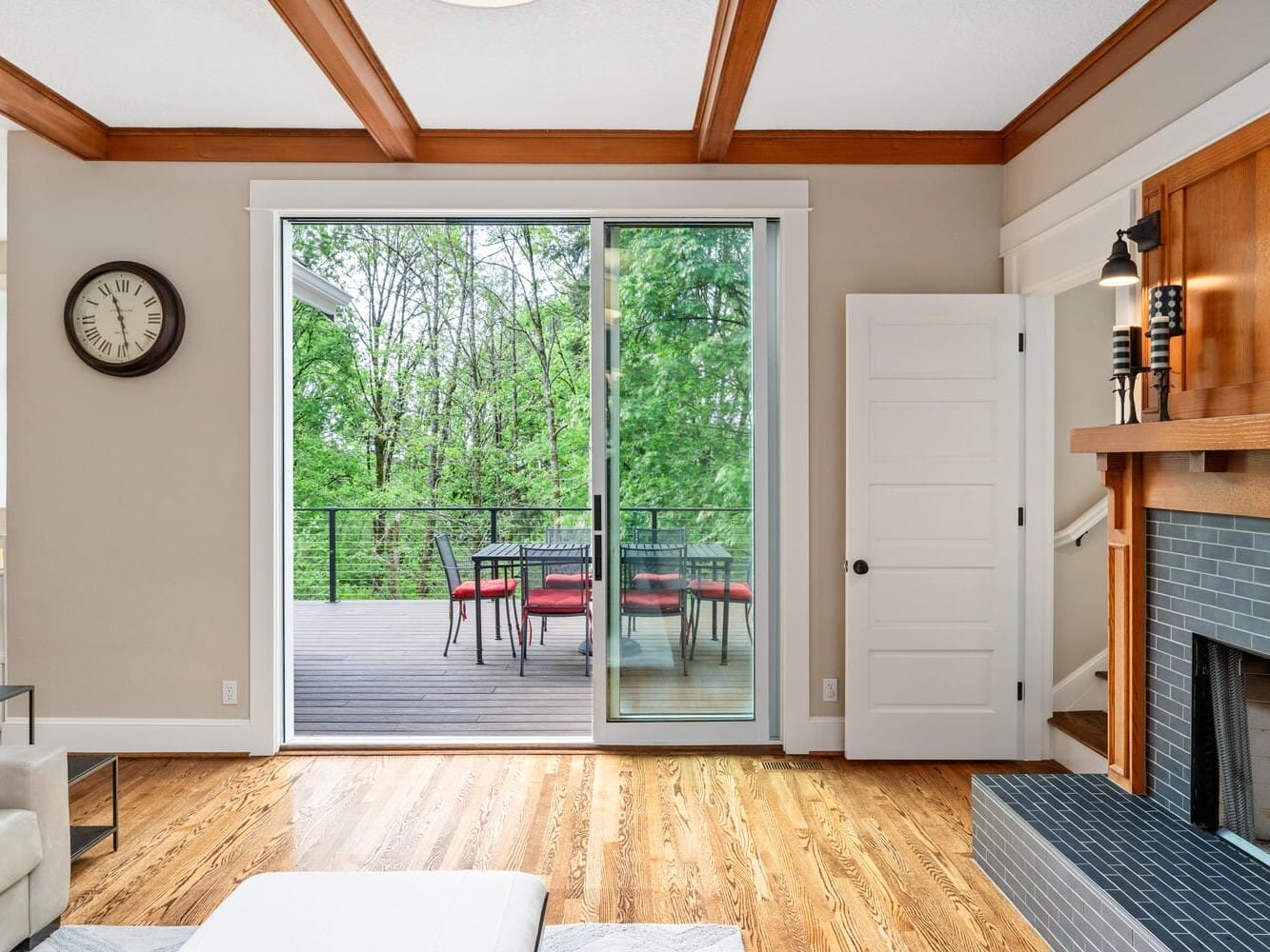 Cozy living room with a sliding glass door leading to a balcony. Inside, there’s a fireplace with blue tiles and wooden mantle, a wall clock, and hardwood floors. Outside, a table with red chairs sits on the patio overlooking green trees.