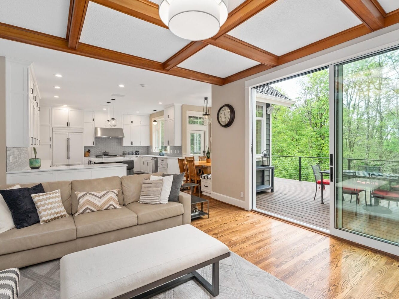 A modern open-concept living area with a beige sectional sofa and patterned pillows, a wooden ceiling with recessed lights, and large sliding glass doors opening to a deck. The kitchen in the background is white with stainless steel appliances.