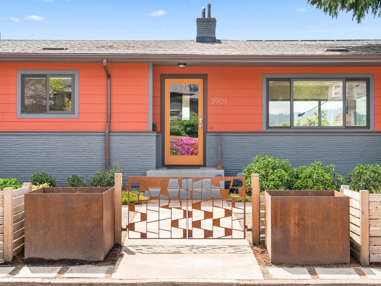 A single-story, modern house with a vibrant orange facade and teal trim. The entrance features a glass door with a house number and is flanked by windows. Geometric metal gate and large planters enhance the landscaped front yard. Clear blue sky above.