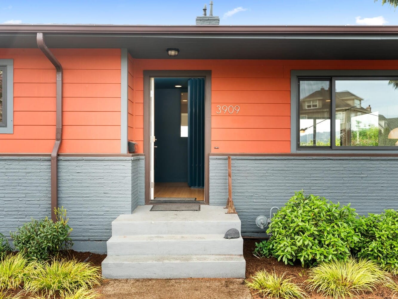 A modern house with orange siding and gray brick accents. The entrance has a small concrete porch with steps leading up. Green shrubs decorate the front, and the house number 3909 is displayed next to the door.