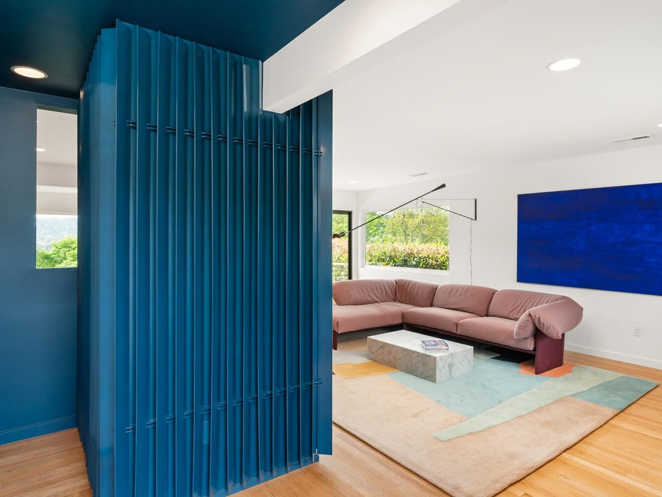 Modern living room with a sleek blue accent wall, a mauve sectional sofa, and a large blue abstract painting. The room features a minimalist design with light wood flooring and a geometric area rug, illuminated by natural light from the windows.