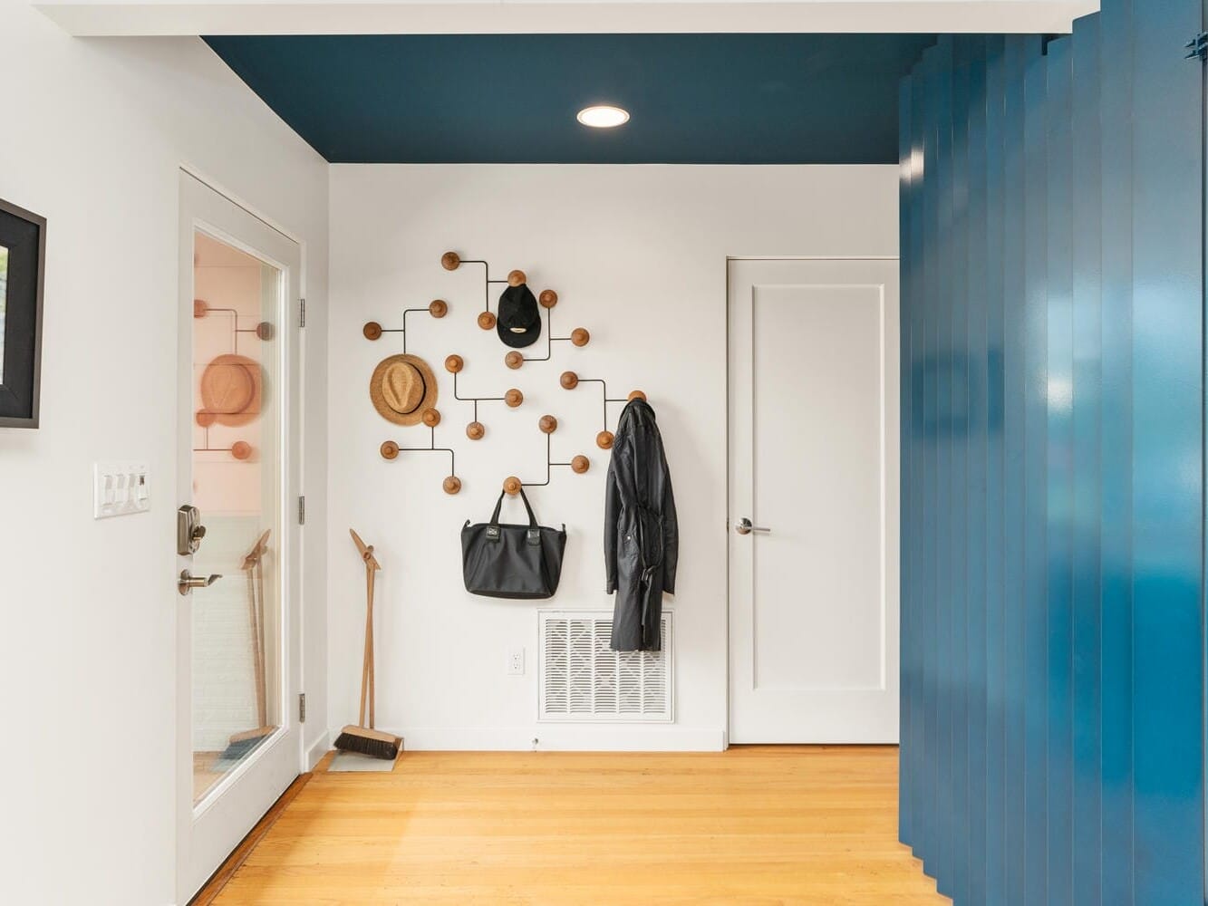 A modern entryway with a light wooden floor, blue accent wall, and ceiling. Wall hooks hold hats, a bag, and a coat. A black broom is in the corner. A framed picture hangs on the white wall, and two doors are visible.