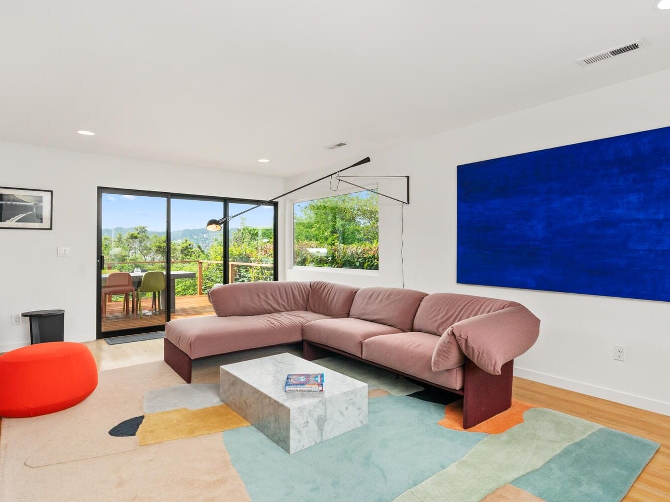 A modern living room with a pink sectional sofa, a white and gray rectangular coffee table, and large blue abstract wall art. The room features a colorful rug and has a view of a patio through glass doors. Vibrant greenery is visible outside.