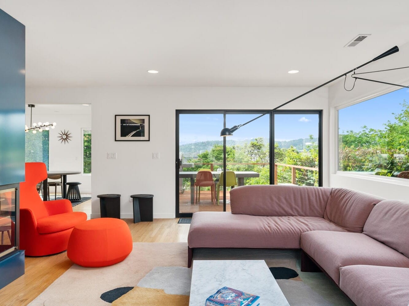 A modern living room with large windows showcasing a scenic view. The room features a pink sectional sofa, a red chair, a white marble coffee table, and a black floor lamp. In the background, theres a dining area and a deck with outdoor seating.