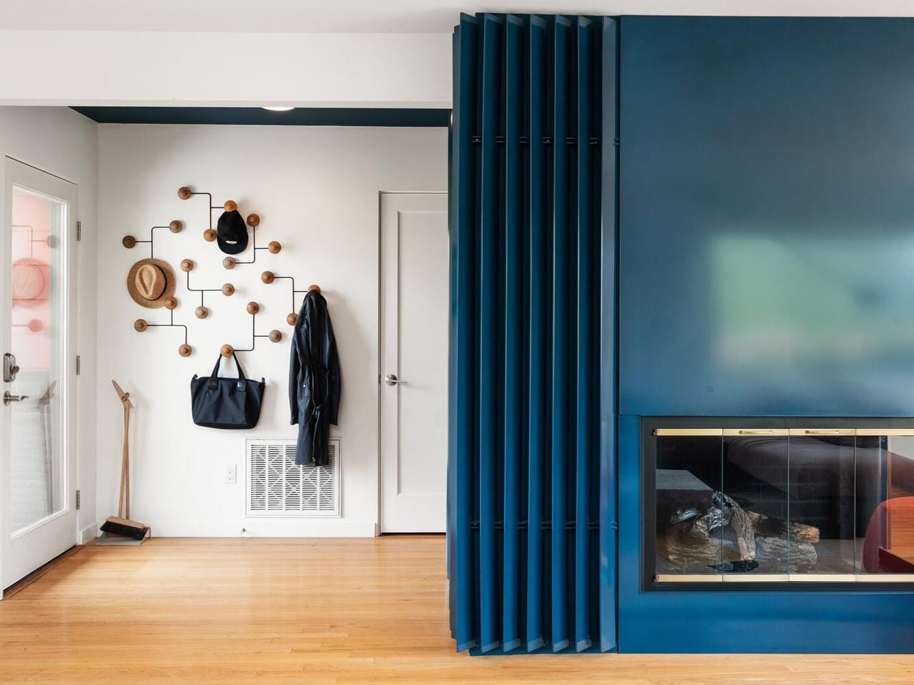 A modern living space featuring a sleek blue accent wall with an integrated fireplace. The entryway has a wall-mounted coat rack with hats, coats, and bags, and a wooden floor contrasts with the contemporary design.