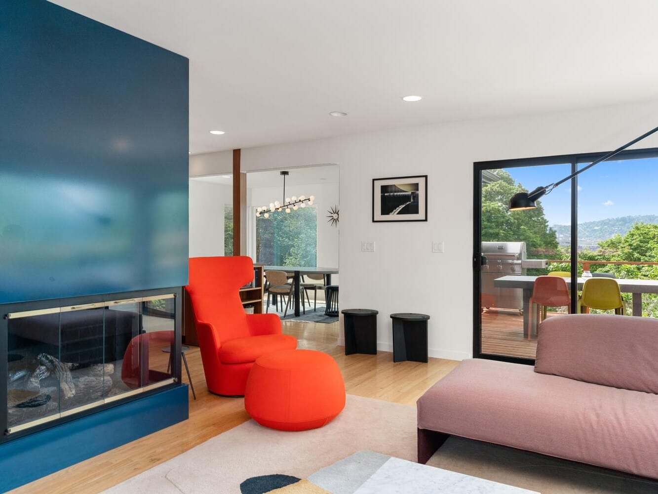 A modern living room with a red armchair and matching ottoman. It features a sleek fireplace, a tan sofa, and a wall-mounted lamp. Large sliding doors open to a balcony with outdoor dining and scenic views of greenery and mountains.