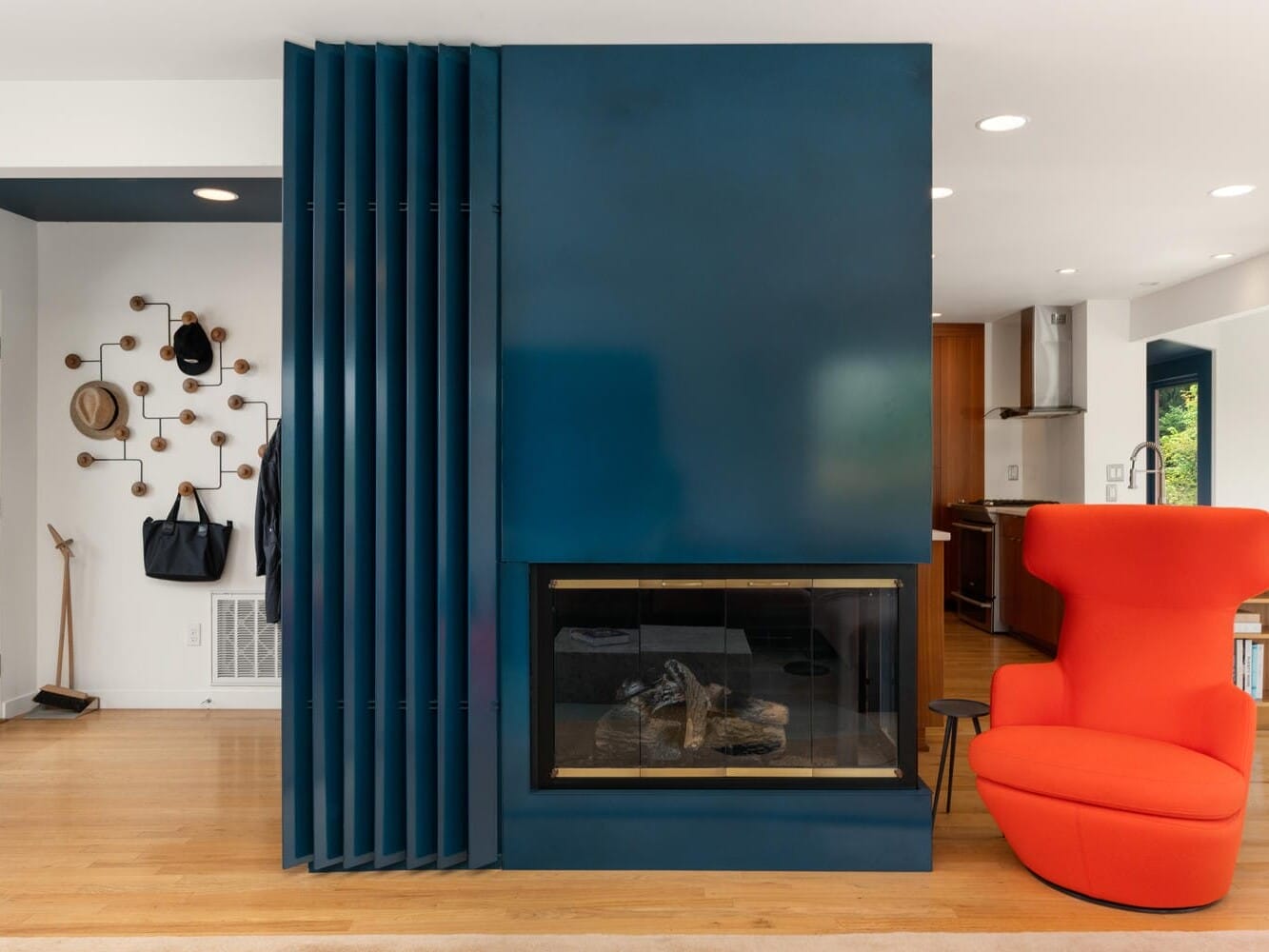 A modern living room features a sleek teal fireplace with a vertical panel design. To the right, a vibrant red, high-back chair sits on a wooden floor. In the background, a wall-mounted coat rack displays hats and bags. The space is airy and contemporary.