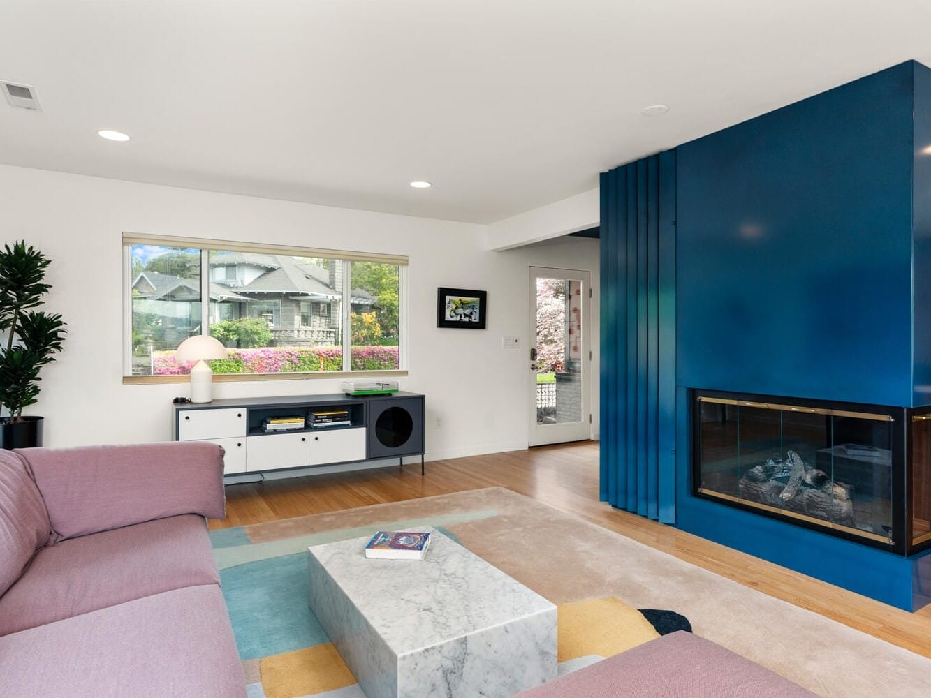 Modern living room with a pink sectional sofa, marble coffee table, and blue accent wall featuring a fireplace. Large window offers natural light and a view of the outdoors. Geometric rug adds a contemporary touch, complementing the minimalist decor.