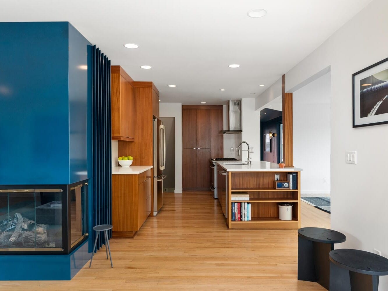 Modern kitchen interior with wooden cabinets and flooring, a blue accent wall, and a glass fireplace. A kitchen island with open shelves holds various items. A framed photo is on the white wall. Two black stools are nearby.