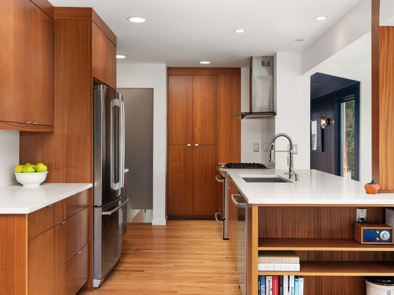 A modern kitchen with wooden cabinets, a stainless steel refrigerator, and a built-in oven. A bowl of green apples sits on the counter. The room has wooden flooring and recessed lighting, with a small bookshelf at the end of the counter.