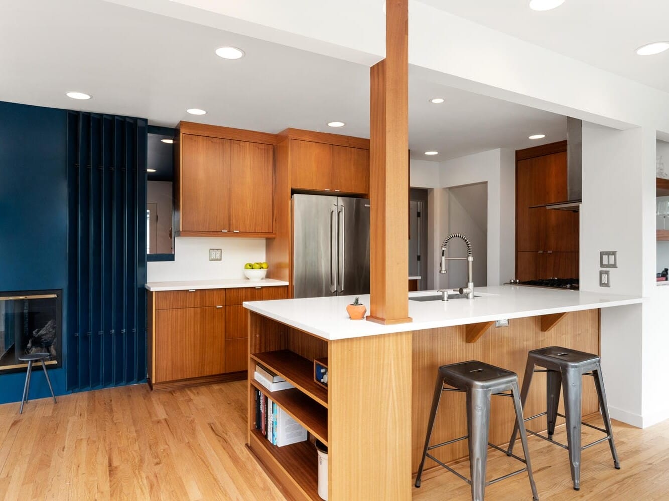 Modern kitchen with wood cabinets, a white countertop island, and stainless steel appliances. Hardwood flooring complements the blue and white walls. Two metal stools are at the island, and a small fireplace is visible. Natural light fills the space.