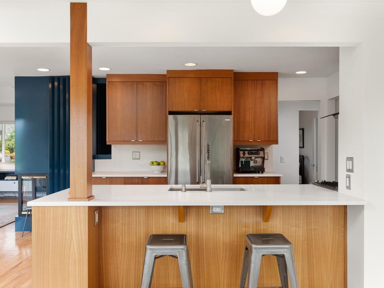 Modern kitchen with wooden cabinets and a large stainless steel fridge. A white countertop with a silver faucet is complemented by two metal stools underneath. In the background, an open living area is partially visible.