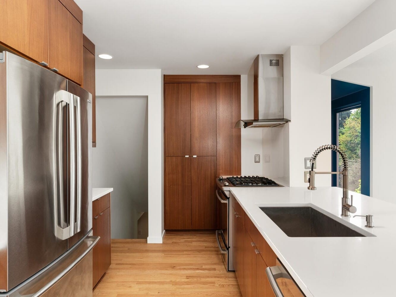 Modern kitchen with stainless steel appliances, including a refrigerator and oven. Features wooden cabinets, a white countertop, and a sink with a silver faucet. Natural light illuminates the space through a window on the right.