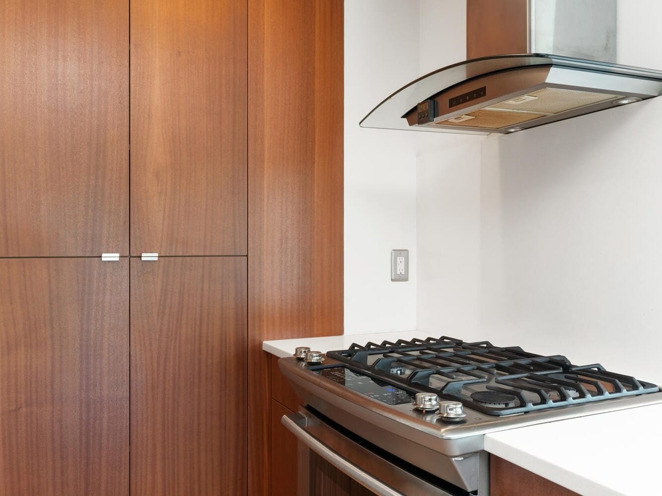 Modern kitchen with a stainless steel stove and range hood. Two large wooden cabinets are to the left of the stove. The countertop is white, giving the kitchen a clean and sleek look.