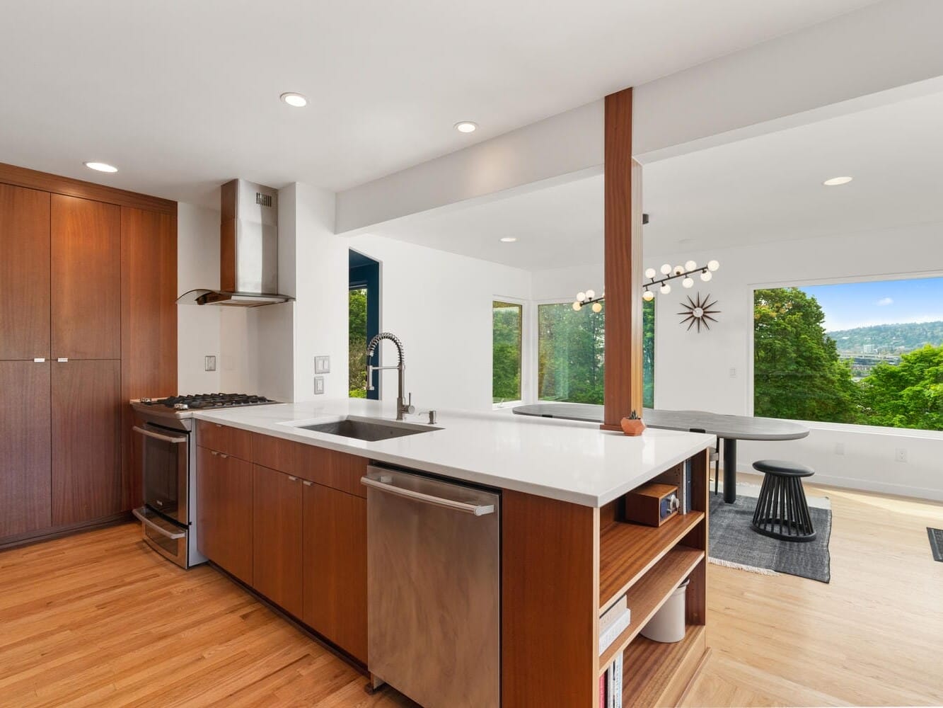 A modern kitchen with wooden floors, white countertops, and stainless steel appliances. The kitchen opens into a dining area with a large window showing lush green trees and a view of distant hills. A unique light fixture hangs over the dining table.