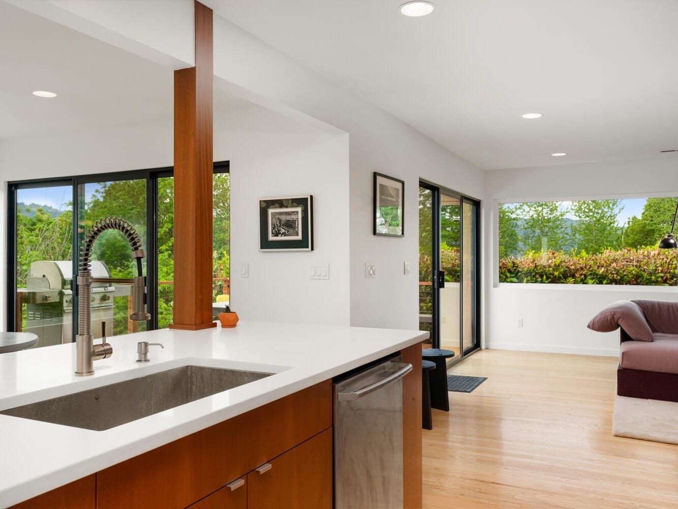 Modern kitchen and living area featuring a large countertop with a sink, sleek faucet, and wooden cabinets. In the background, theres a cozy sofa and large windows offering views of greenery and natural light.