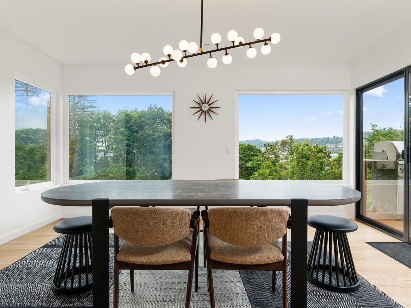 A modern dining room with a long oval table, surrounded by four brown chairs. There are large windows with views of lush green trees. A stylish chandelier hangs above, and a decorative starburst clock is on the white wall. There is an outdoor grill visible.