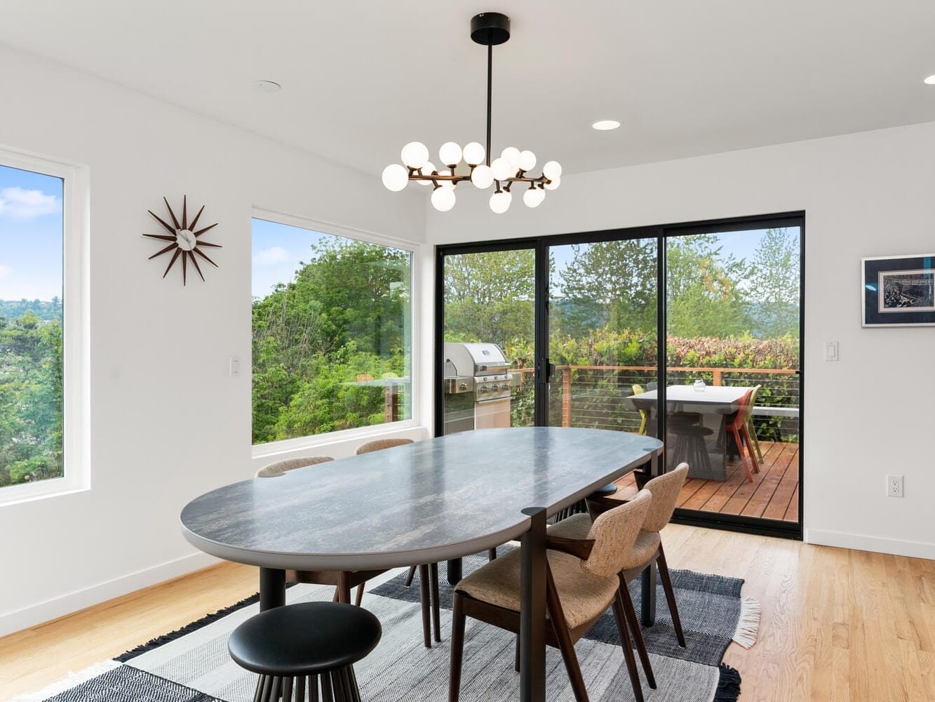 A modern dining room with a dark oval table and four chairs. Large windows and sliding doors let in natural light and offer views of a scenic landscape. A chandelier hangs above the table, and a wooden deck with a grill is visible outside.