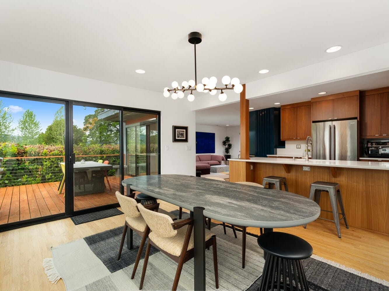 A modern open-plan dining and kitchen area with a gray oval table, beige cushioned chairs, and a contemporary light fixture. The kitchen features wood cabinets and stainless steel appliances. Glass doors lead to a wooden deck with outdoor seating and lush greenery.